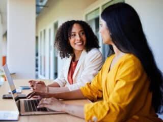 Two diverse investors talking and smiling while working on a laptop together at a table in an office lounge